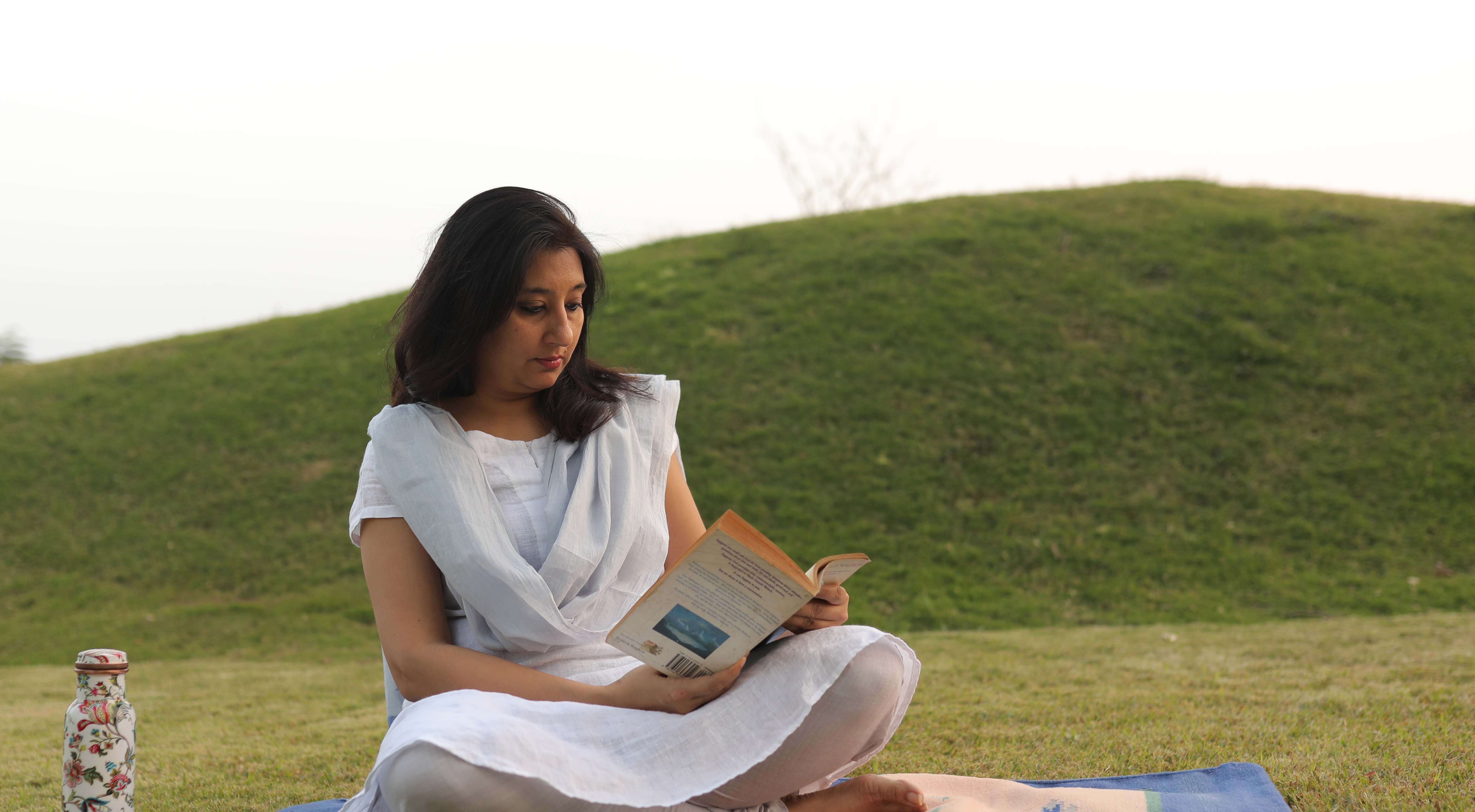 Ishita seated cross-legged on a blanket in a grassy field, reading a book under an open sky.